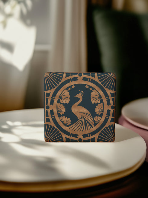 Square ceramic coaster featuring an Art Deco copper peacock design centered in a medallion, surrounded by fan patterns and shell motifs on a dark forest green background, with cork backing.